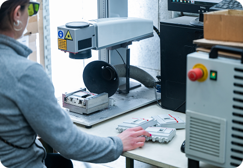 Operatrice con occhiali di protezione utilizza una macchina laser per incisione su componenti di plastica. È seduta a un banco di lavoro con pezzi già lavorati accanto a lei. Il contesto suggerisce un processo di marcatura industriale in un ambiente di produzione tecnologico.