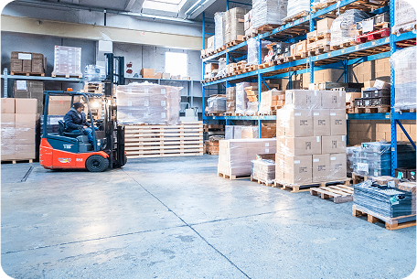 Magazzino industriale con scaffalature cariche di scatole e pallet. Un operatore manovra un carrello elevatore rosso per spostare la merce. L’ambiente è ben illuminato e organizzato, suggerendo un'attività di stoccaggio e logistica per la gestione dei materiali.